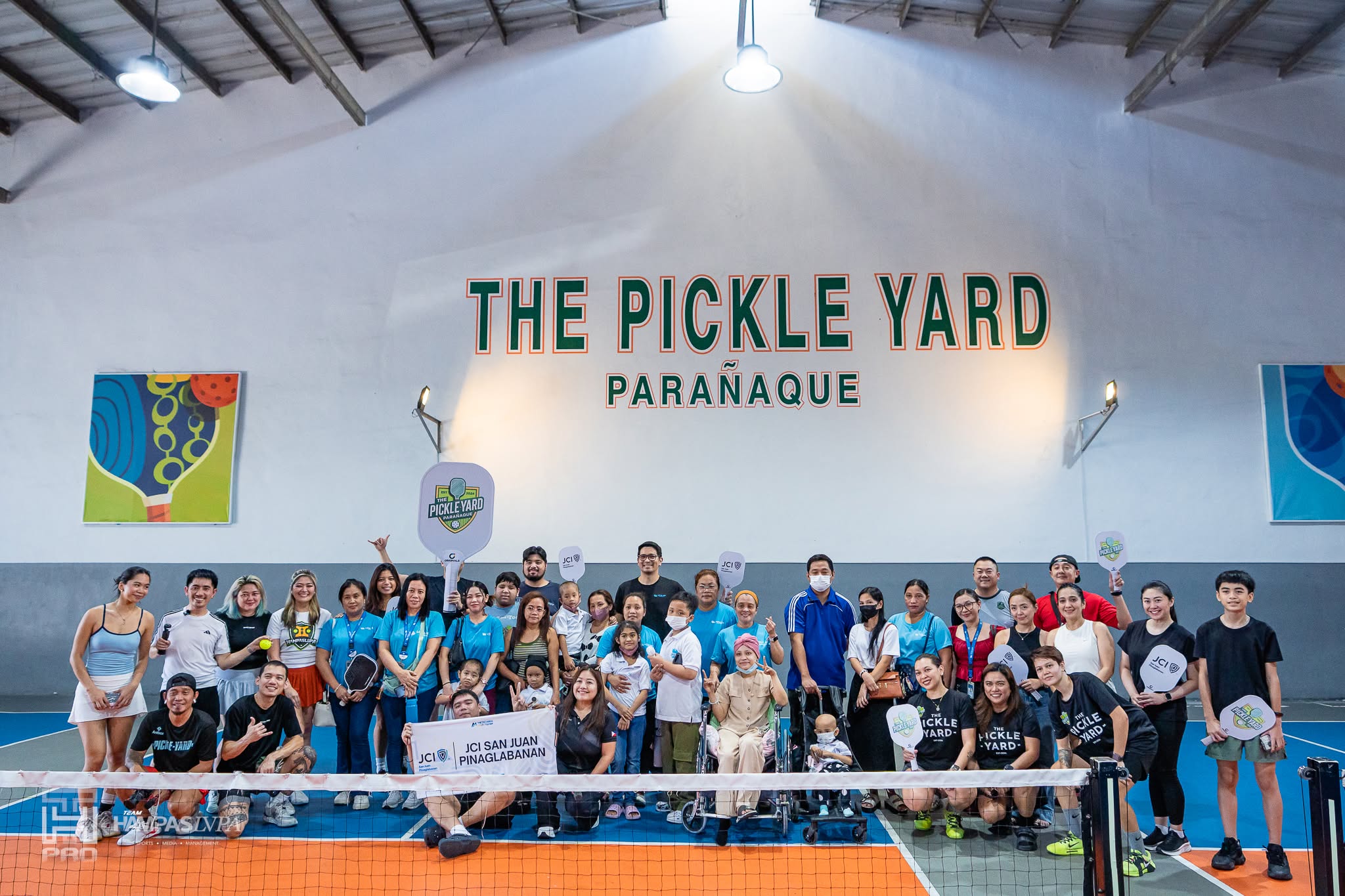 A large group of players and guests posing on court beneath a painted The Pickle Yard Parañaque sign